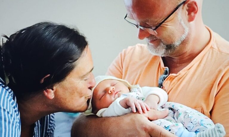 Eine Frau küsst ein schlafendes Baby in den Armen eines lächelnden Mannes mit Brille. Jörg Tuschling - Interim Operations Management