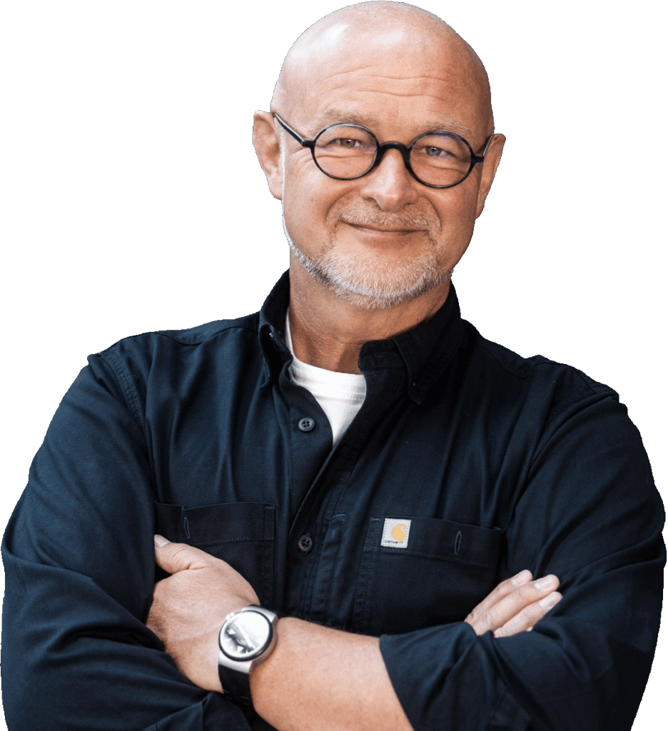 Ein glatzköpfiger Mann mit Brille und gestutztem Bart, der ein schwarzes Hemd und eine Armbanduhr trägt, steht mit verschränkten Armen und lächelt in die Kamera. Jörg Tuschling - Interim Operations Management